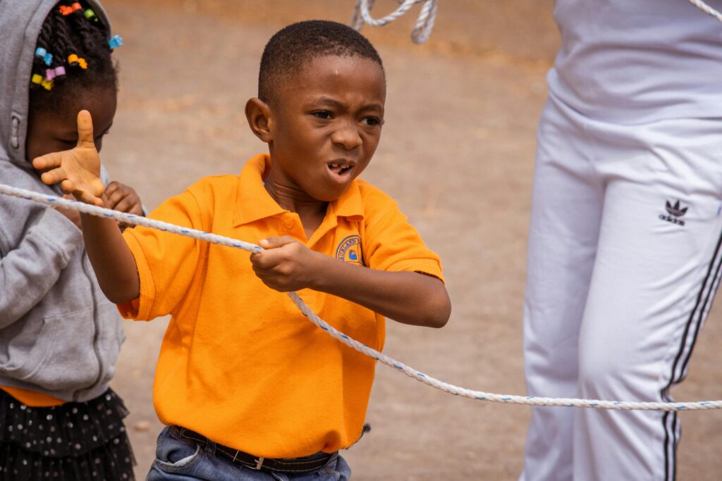 A young boy in an orange shirt enthusiastically pulls on a rope during an outdoor tug of war competition.
