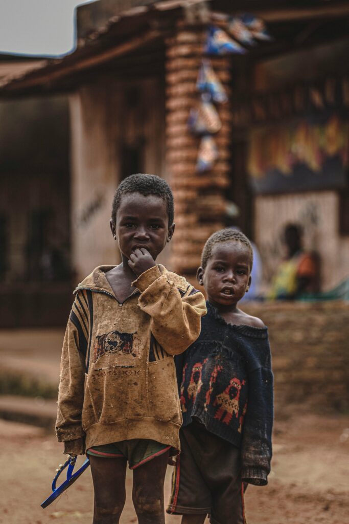 Two young boys in worn clothing standing outdoors, looking somber and pensive.